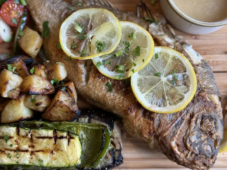 Whole roasted fish topped with lemon slices beside grilled vegetables